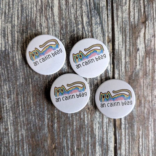 badges with a colorful cat design and text 'an caitin beag' on a wooden surface.